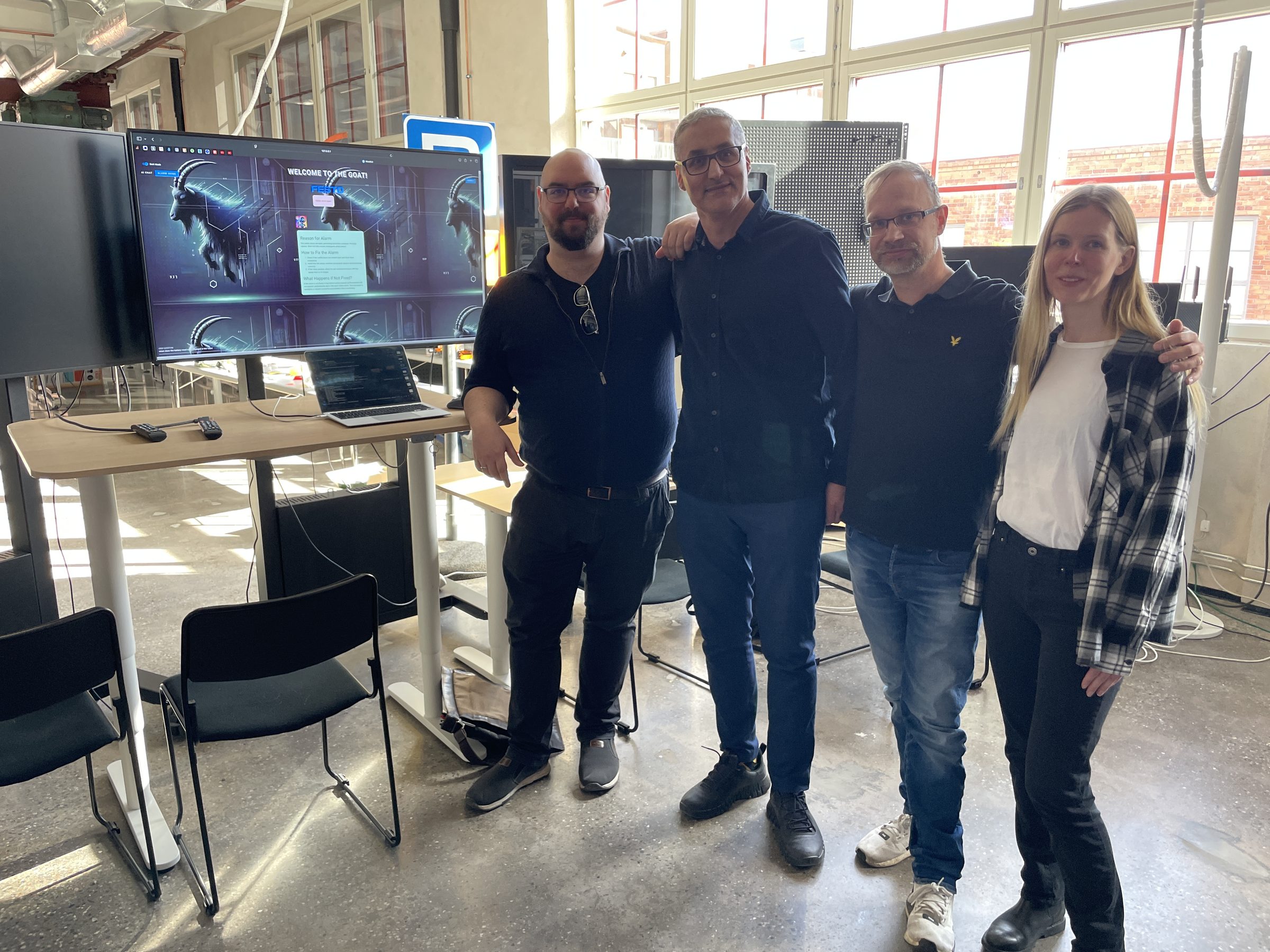Four people standing in front of a large display running the Talking Systems assistant at the testbed